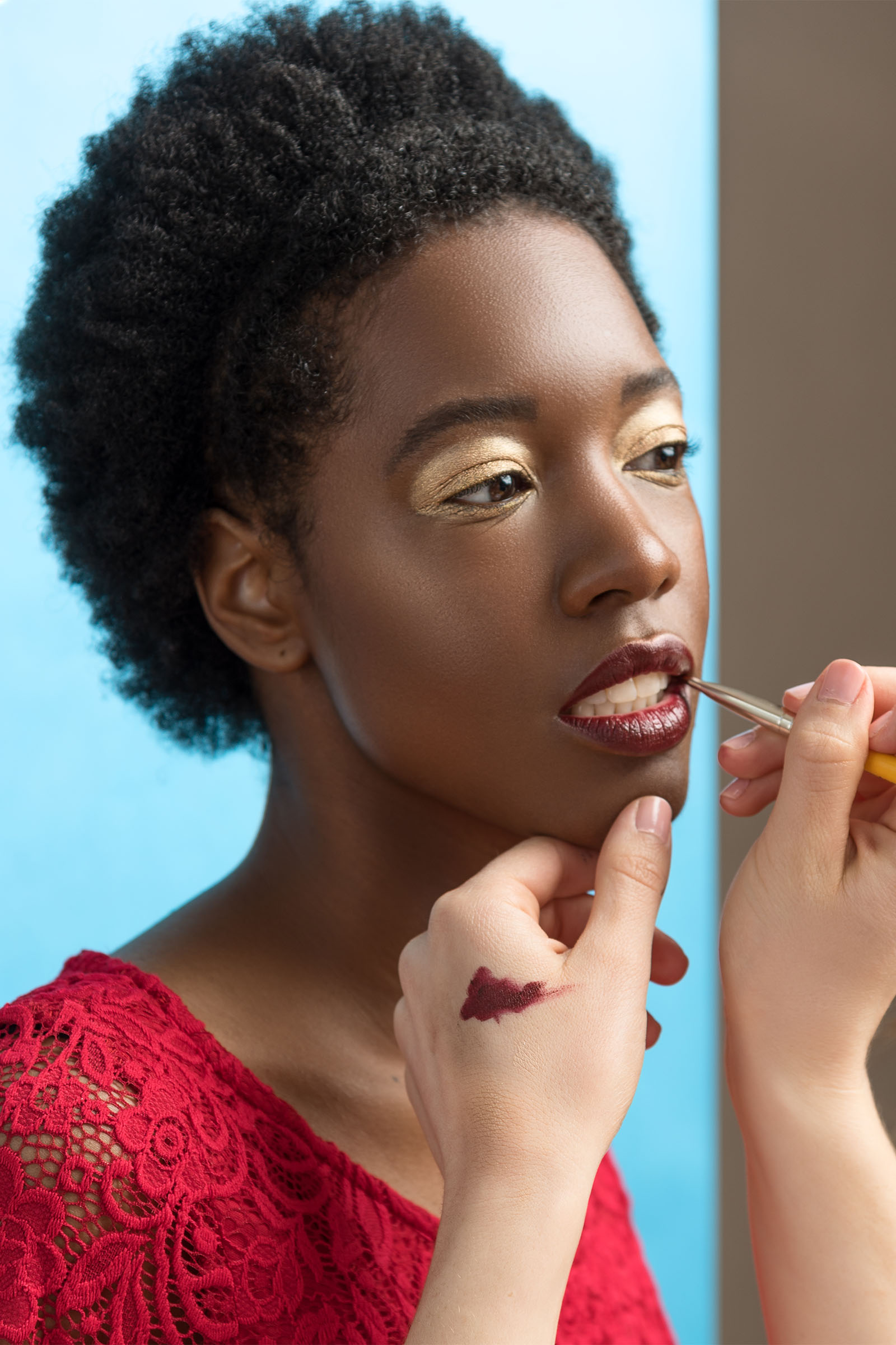 Beautiful Black female model with golden eye shadow, makeup artist is applying lipstick on her lips, baby blue background by Utah photographer Christopher Loke